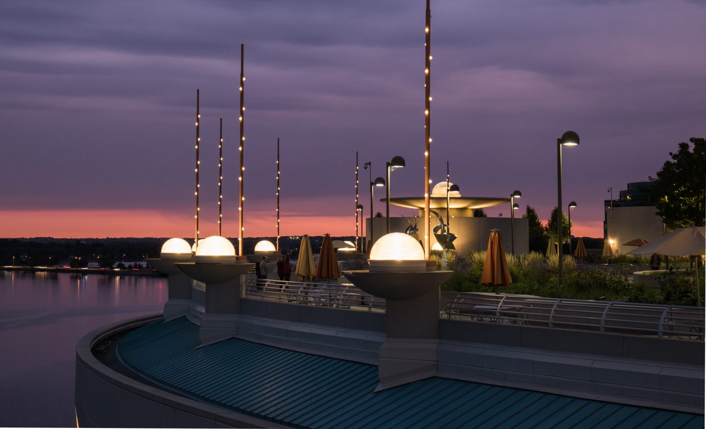 The Monona Terrace rooftop gardens at dusk, with lit globe lamps, patio umbrellas and Lake Monona under a purple sunset sky.